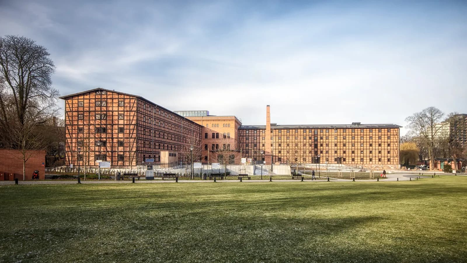 A large, multi-story brick building with evenly spaced windows and a half-timbered facade, partially obscured by a tree and surrounded by a grassy lawn under a partly cloudy sky.