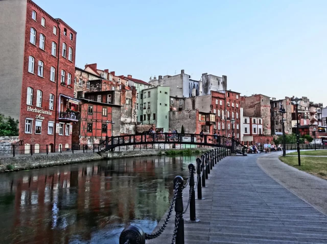 A scenic view of a river with a small arched bridge, flanked by old colorful buildings on one side and a paved promenade with a chain railing and park benches along a grassy area on the other.