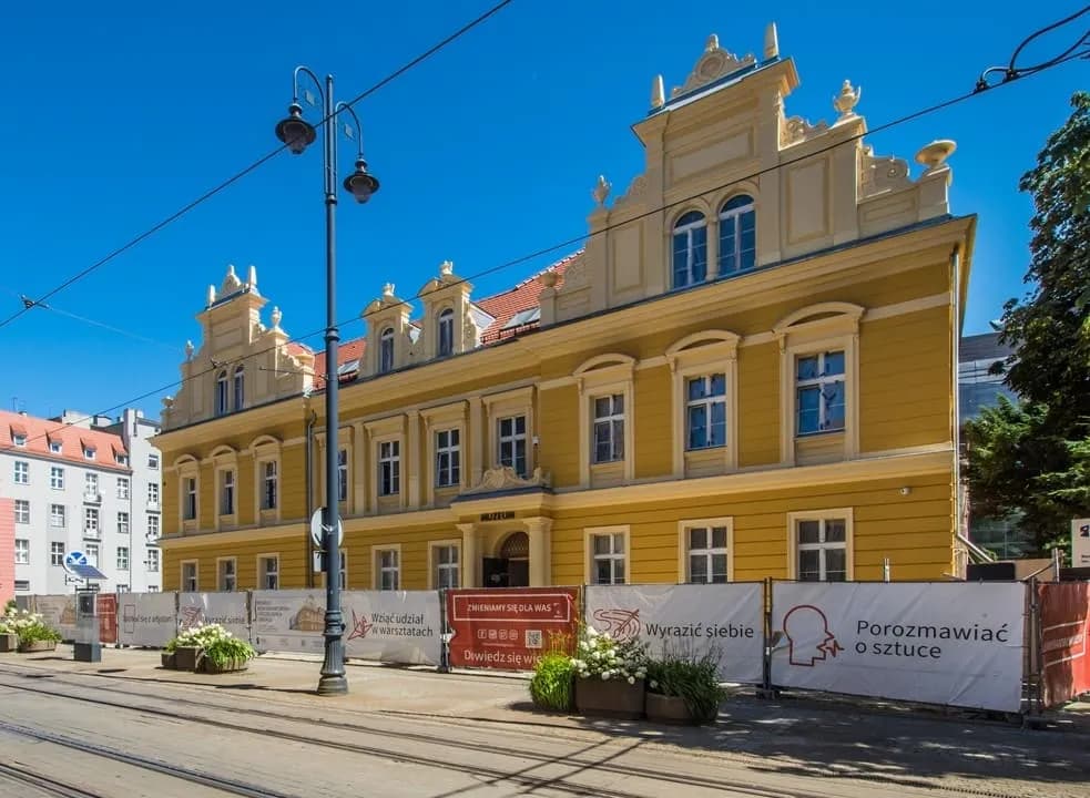 A grand, multi-story yellow building with intricate architectural details, including ornate gables and numerous windows, stands against a clear blue sky, with a lamppost and construction barriers with signs in the foreground.